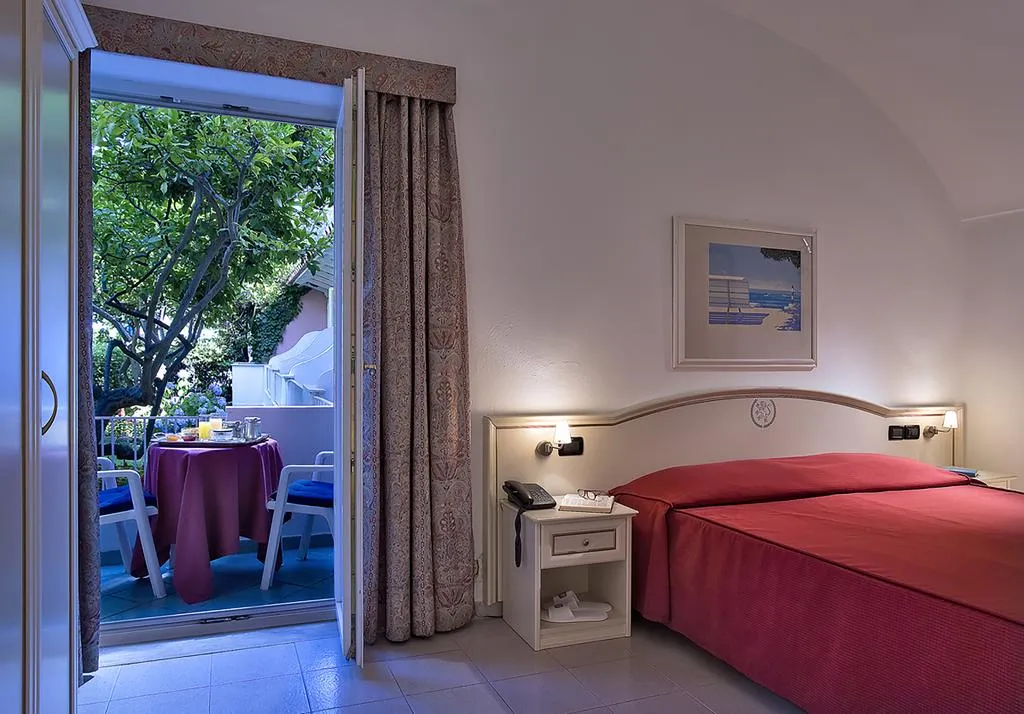 Camera con letto matrimoniale dal copriletto rosso e uscita su balcone con tavolino per la colazione nel verde